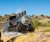 A vintage steam locomotive is chugging along a track through a dry hilly landscape under a clear blue sky