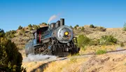 A vintage steam locomotive is chugging along a track through a dry, hilly landscape under a clear blue sky.