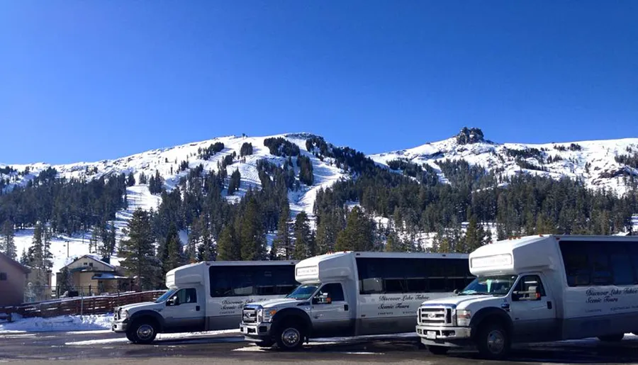 Two shuttle buses are parked at the base of a snow-covered mountain under a clear blue sky.