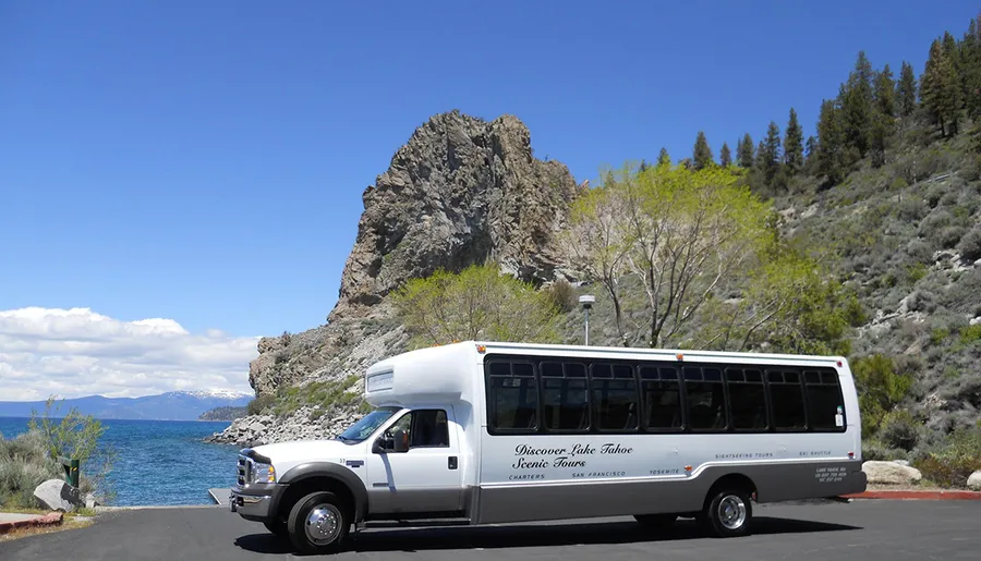 A tour bus promoting scenic tours of Lake Tahoe is parked beside a lake with a rugged mountain in the background under a clear blue sky.