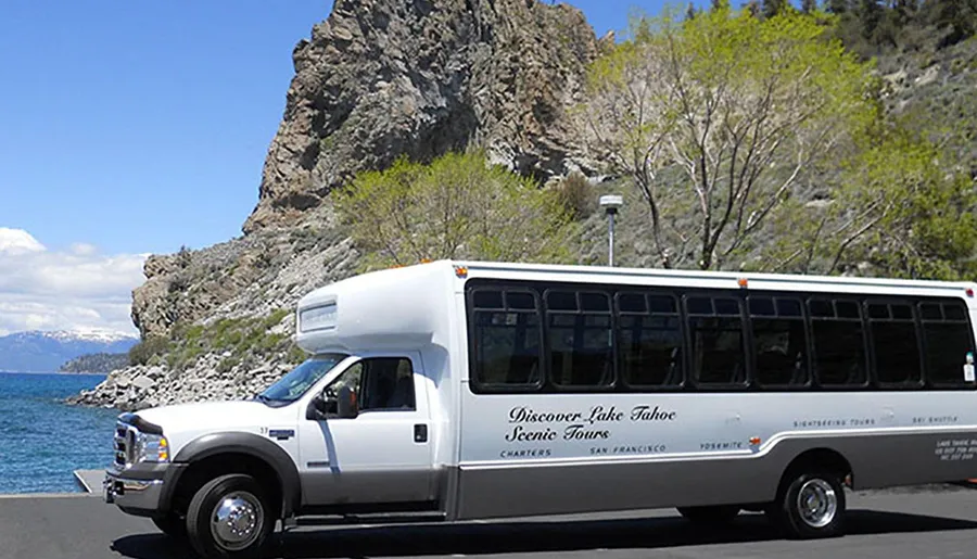 A tour bus with Discover Lake Tahoe Scenic Tours printed on its side is parked near a lakeshore with a rocky outcrop in the background under a clear blue sky.