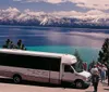 A group of tourists stands beside a scenic tours bus admiring the stunning view of a snow-capped mountain range across the vibrant blue waters of a lake