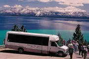 A group of tourists stands beside a scenic tours bus, admiring the stunning view of a snow-capped mountain range across the vibrant blue waters of a lake.