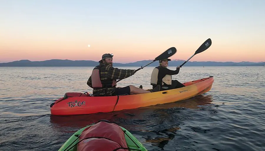 Two people in a tandem kayak are paddling on calm waters with the sunset and a full moon in the background.