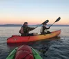Two people are paddling a tandem kayak on tranquil waters at sunset with a beautiful sky gradient in the background and a view of the tops of other kayaks in the foreground