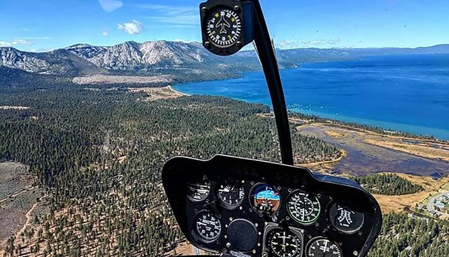 The image shows the view from a helicopter cockpit overlooking a scenic landscape featuring a large lake, surrounded by forests and mountains.