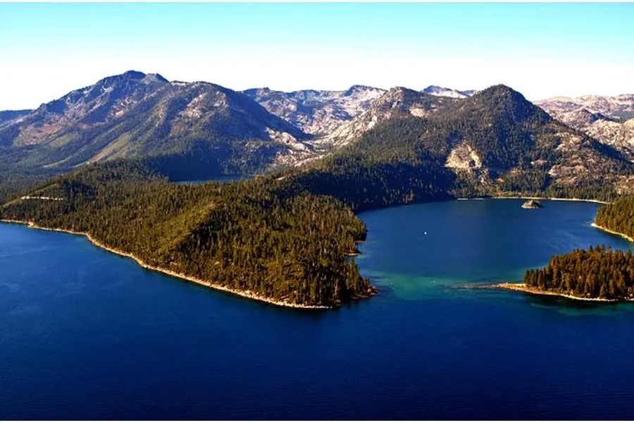 The image shows a breathtaking aerial view of a large, forest-covered peninsula jutting into a serene lake with mountains in the background.