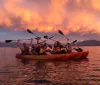 People in colorful kayaks enjoy a serene paddle on calm water under a dramatic orange sunset sky