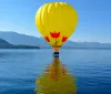 A bright yellow hot air balloon is floating above a serene lake with its reflection on the water and a backdrop of mountains under a clear blue sky