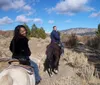 Two individuals are seen horseback riding in a scenic landscape with sparse vegetation under a partly cloudy sky