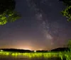 A starry night sky stretches above a serene lake with a small island and distant lights from a lakeside community