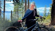 A person is standing with a bicycle in a forested area, smiling at the camera, with a lake visible in the background.