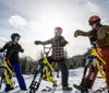 Three people equipped with helmets are standing in the snow each on a yellow snow bike with the sun setting in the background