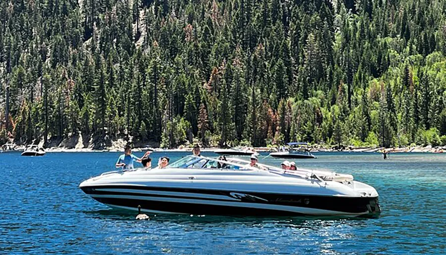 A group of people enjoys a sunny day on a motorboat on a clear blue lake, surrounded by a dense pine forest.