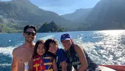 A family is smiling and enjoying a sunny day on a boat with a scenic mountainous backdrop.