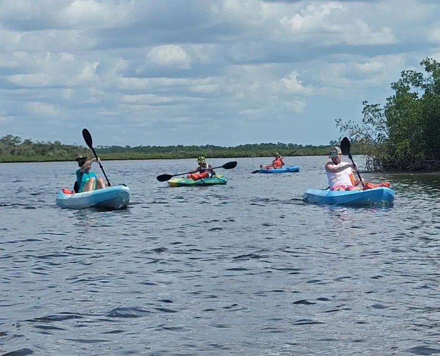 A group of people are kayaking on a calm river flanked by greenery under a partly cloudy sky.