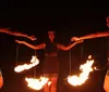 Performers are engaged in a dynamic fire dance possibly part of a cultural performance with a man in the center twirling fire sticks against a dramatic fiery backdrop