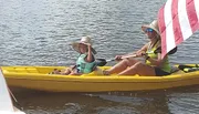 Two individuals, one adult and one child, are paddling on a yellow kayak with an American flag-themed sail attached.