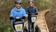 Two people wearing helmets are smiling at the camera while standing on Segways on a wooden path through a forested area.