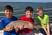 Three smiling young boys are on a boat, holding a large fish together against the backdrop of a bright, sunny sky and water.