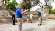 A guide wearing a blue shirt is gesturing and speaking to a small group of visitors at an outdoor historical site with stone structures and trees.