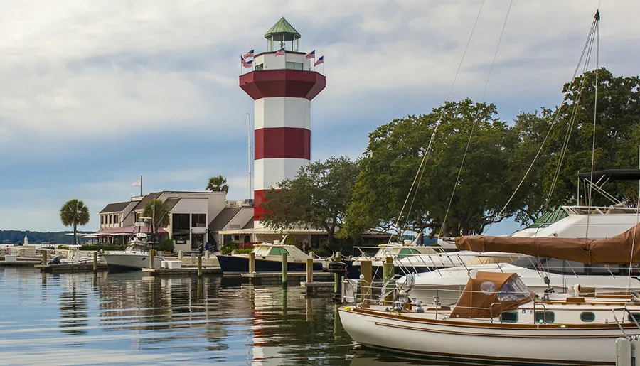 The image shows a picturesque marina with boats docked in calm waters, and a distinctive red and white lighthouse standing prominently in the background.