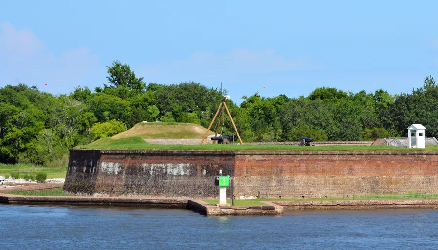 The image shows a historic fortification with grassy embankments and antique cannons overlooking a body of water, under a clear blue sky.