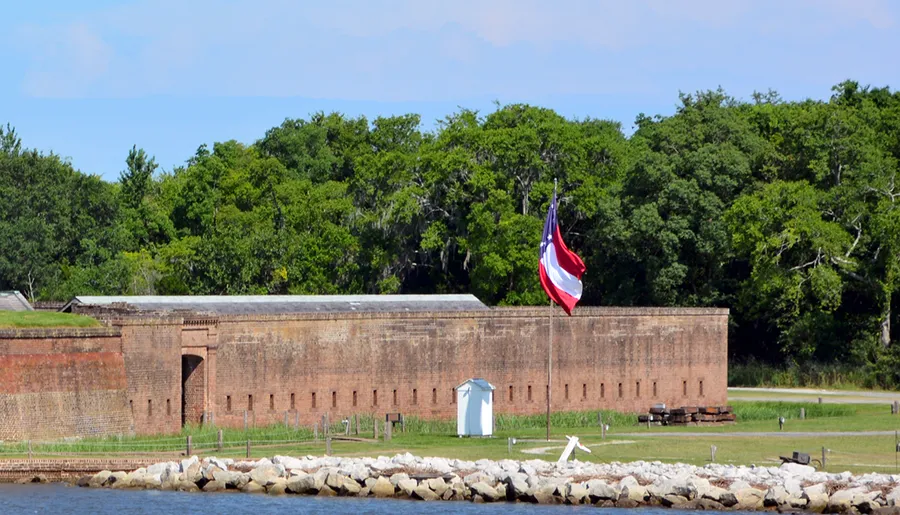 An American flag flies prominently over a historic brick fortification surrounded by greenery and a stone shoreline.