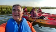 A group of smiling people in life jackets are enjoying a sunny day while kayaking on calm waters surrounded by tall reeds.