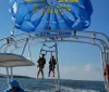Two people are preparing to parasail over the water with a vibrant blue parasail emblazoned with Hilton Head Harbor as they are tethered to a boat with a clear sky in the background