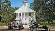 Visitors in golf carts are parked in front of a quaint white church surrounded by greenery under a blue sky.