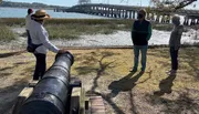 Three people are standing near an old cannon with a scenic water view and a bridge in the background.