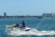 A person is riding a jet ski on a bright day, with a city coastline visible in the background.
