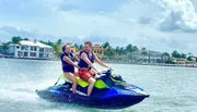 Two people are enjoying a jet ski ride on a sunny day with waterfront houses in the background.