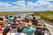 A group of tourists is enjoying an airboat tour through a grass-lined waterway under a clear blue sky.