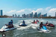 A group of people are riding jet skis on a body of water with a city skyline in the background.