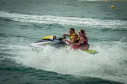 Two people are riding a yellow and purple jet ski over choppy waters, creating a spray of water behind them.