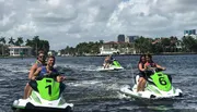 Three pairs of people are riding jet skis on a waterway, with waterfront houses and buildings in the background.