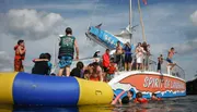 People in life jackets enjoy water activities around a partially submerged inflatable raft with onlookers capturing the moment from a boat named 