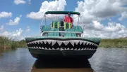 A person is standing on a pontoon boat designed to look like an alligator with its jaws open, floating on calm water under a partly cloudy sky.