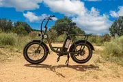An electric bicycle is parked on a dirt path with a backdrop of blue sky and scattered clouds, surrounded by brush and trees.