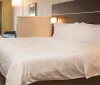 The image displays a neatly made hotel room bed with white linens in a modernly furnished room with a seating area near the window