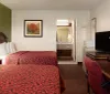 The image shows a standard hotel room with two double beds with red patterned bedspreads a nightstand with a lamp a work desk with a television on it and an open bathroom door revealing a sink and mirror