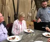 A chef is presenting a dish to two guests seated at an outdoor dining table with wine glasses and plates of food