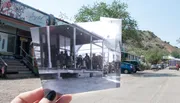 A person's hand is holding up a black and white photo of an old post office, blending the past scene with the current street view in color.