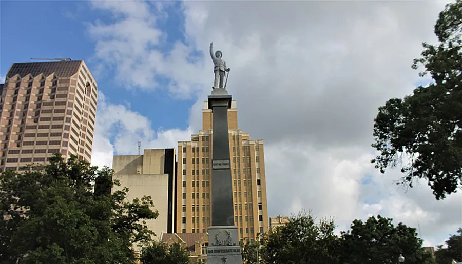 The image shows a tall monument with a statue on top, set against a backdrop of modern buildings and a cloud-filled sky.