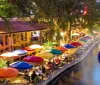 An outdoor riverside dining area illuminated at night with vibrant colorful umbrellas and people enjoying their evening