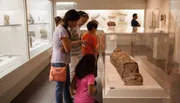 Visitors are closely examining ancient artifacts at a museum exhibit, with a focus on a sarcophagus.