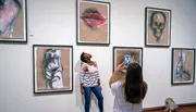 A person admires artwork in a gallery while another takes a photo with their smartphone.