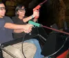Two people are smiling and wearing 3D glasses seemingly enjoying a ride or attraction where they hold a toy gun together against a backdrop that resembles a stone tunnel